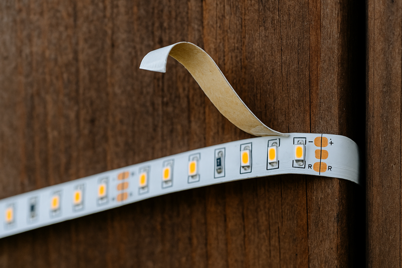 An LED strip light peeling away from a wooden fence, showing failed adhesive