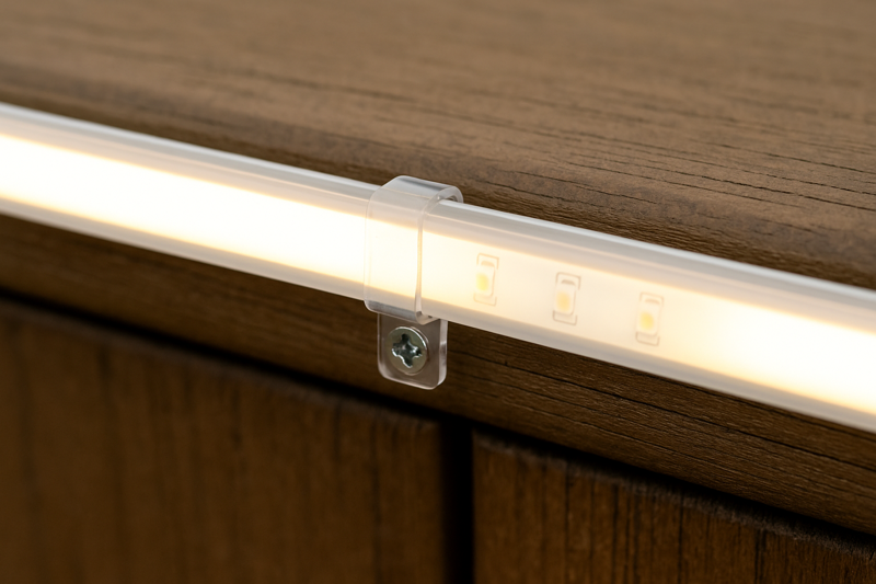 A close-up of a clear plastic mounting clip screwed into a wooden deck, securely holding an outdoor LED strip in place.