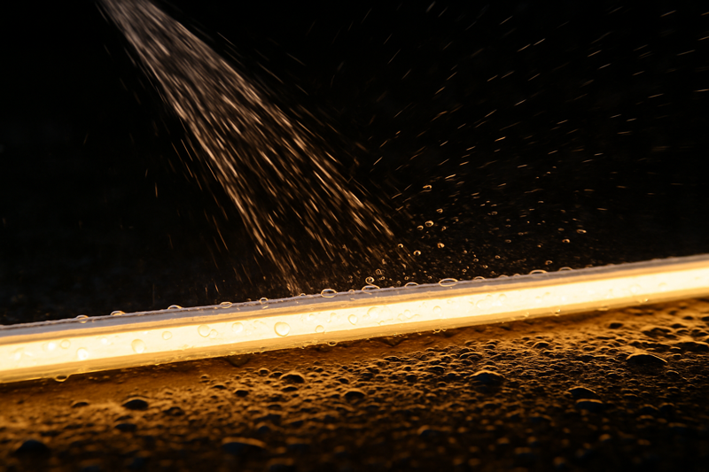 A close-up shot of water from a sprinkler splashing onto a glowing IP65-rated LED strip, showing the water beading up on the silicone coating.