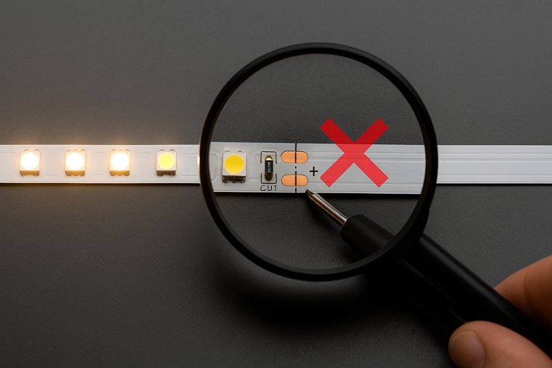 An image of an LED strip with the first 3-LED segment lit up brightly, and the remainder of the strip completely dark. A magnifying glass is focused on the cut line just after the lit segment.
