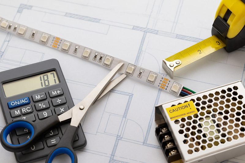 A styled photo showing an LED strip, a calculator with numbers on it, a tape measure, and a power supply, neatly arranged on a blueprint.