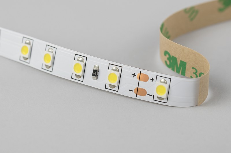 A close-up, high-detail shot of a flexible LED strip, showing the individual LED chips, the circuit board, and the peel-and-stick adhesive backing.