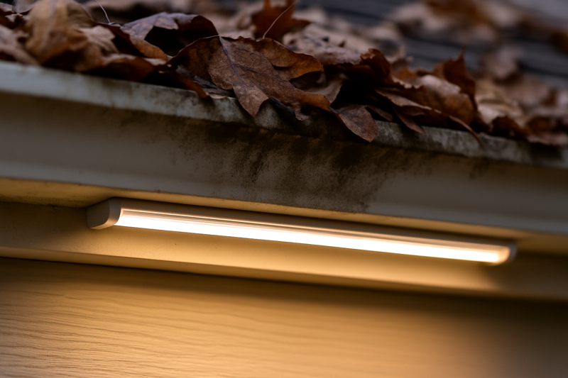A close-up of a dirty gutter area filled with wet brown leaves. Just below it, under the soffit, is a clean, pristine LED strip inside a covered aluminum channel, glowing brightly, unaffected by the mess above.