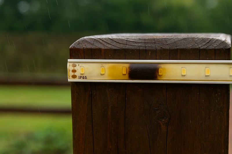 A photo showing an IP65 epoxy-coated strip installed on an exposed fence post, with visible yellowing and a section that has failed and gone dark after a rainstorm.