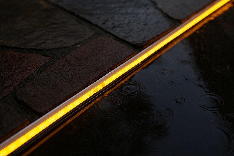 A vibrant IP67 LED strip installed along a stone pathway, partially covered in a puddle after a rain shower, glowing brightly and operating perfectly.