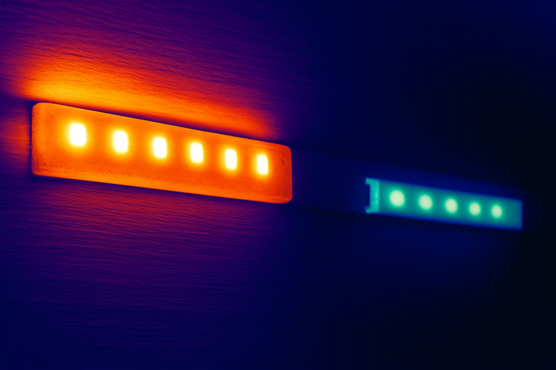 A thermal camera image showing two strips. The one on the left, stuck directly to wood, is glowing hot orange and red. The one on the right, in an aluminum channel, is cool blue and green.