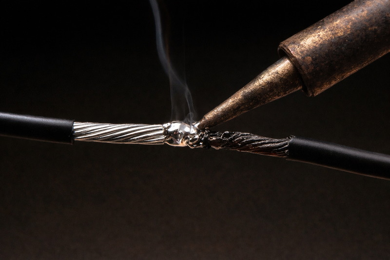 Close up of two wires. One is bright silver (tinned), the other is corroded black copper. A soldering iron is sealing a connection.