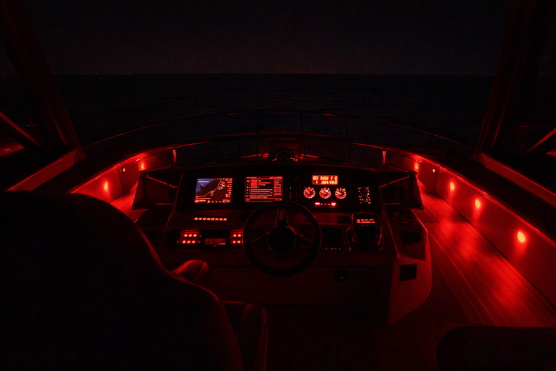 A boat captain's view from the helm at night. The instrument panel and floor are lit with dim red light, allowing a clear view of the dark ocean outside.