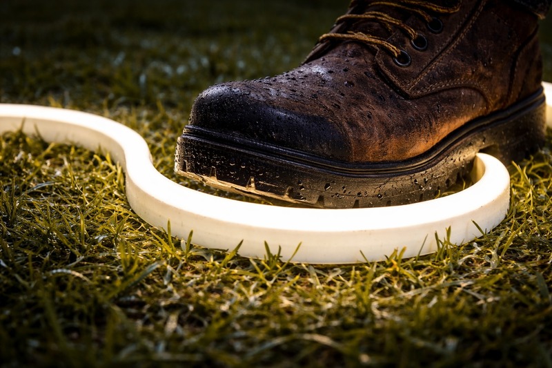 Close up of a thick, flexible white silicone neon strip resting on wet grass, with a heavy boot stepping on it to demonstrate durability.