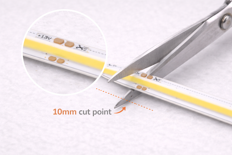 Close up photo: A pair of scissors cutting a 12V COB strip at a tiny mark labeled 